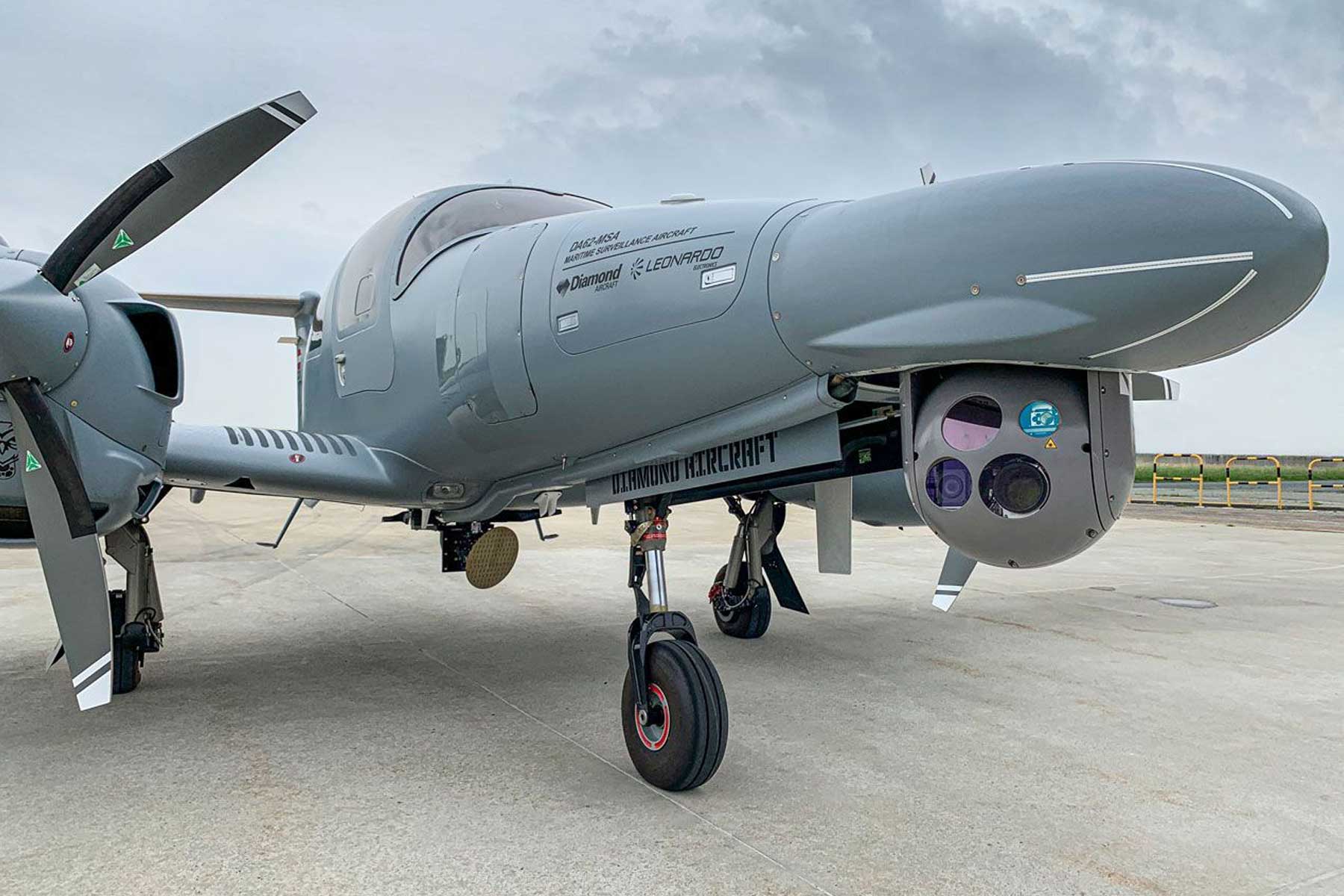 Gray surveillance aircraft on a concrete surface, equipped with sensors and a propeller, under a cloudy sky.