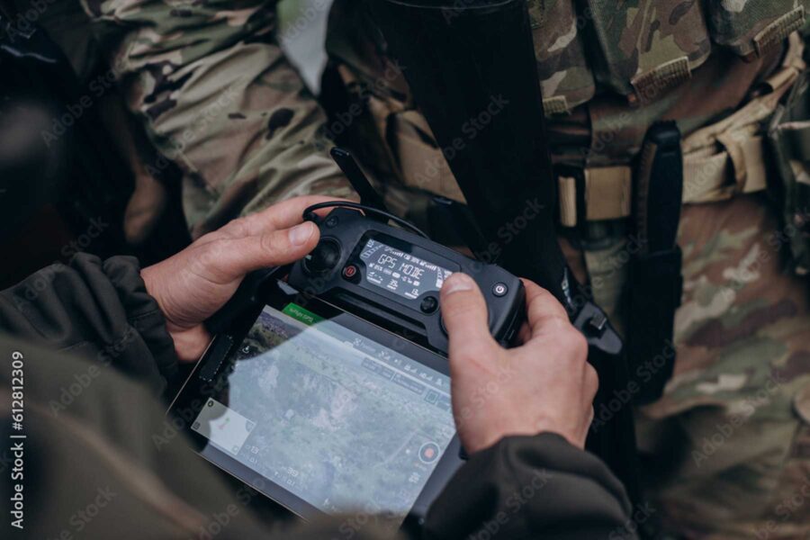 Person using a drone controller with a screen displaying a map. Two individuals wearing camouflage gear are visible in the background.