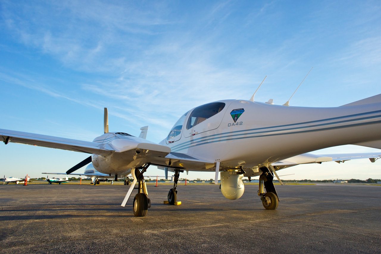 Small white twin-engine airplane parked on an airfield under a clear blue sky.