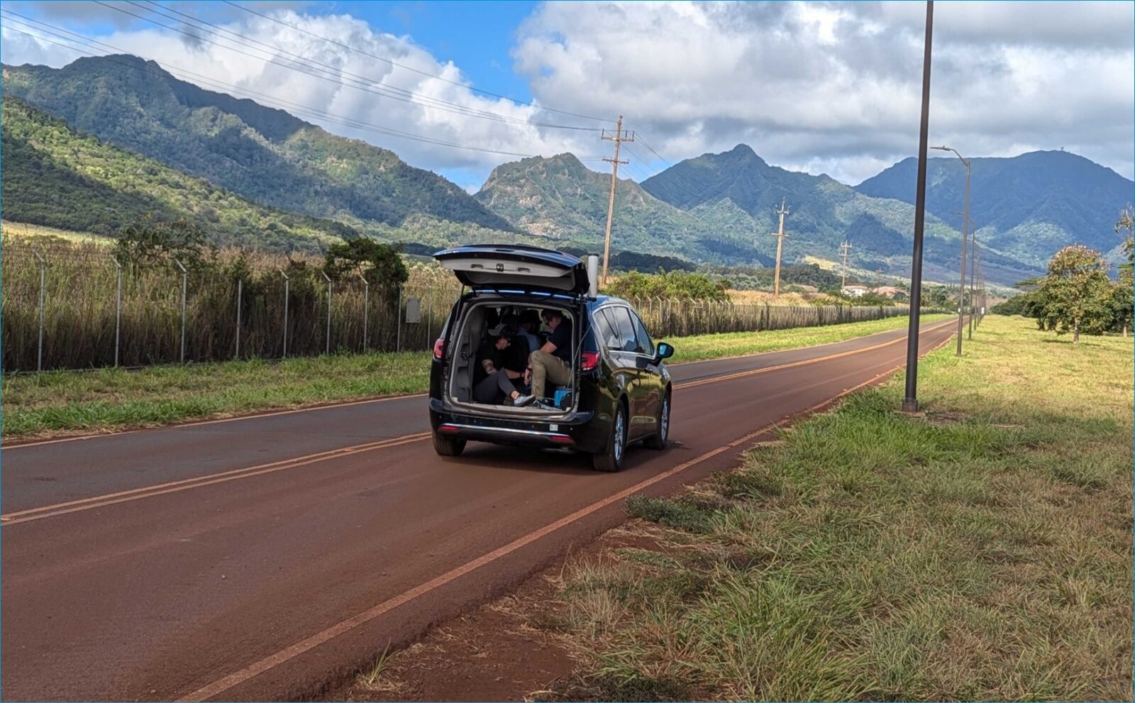 Minivan with open trunk parked on a rural road. Two people are sitting inside, with a view of grass, trees, and hills in the background.