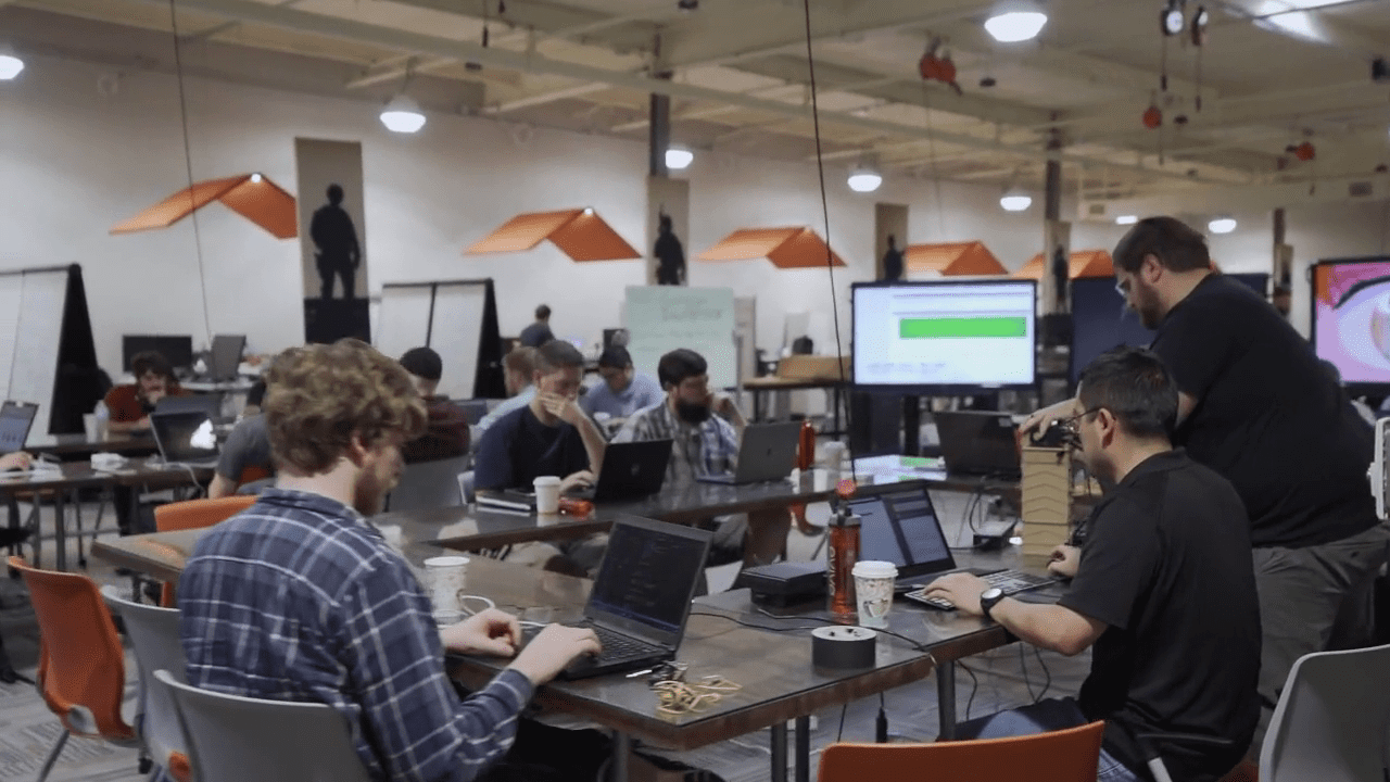 People working on laptops in a spacious office with an open layout. Various tables are arranged around the room with multiple individuals engaged in focused activities.