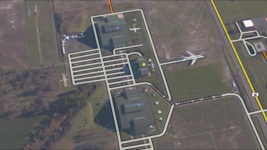 Aerial view of museum buildings with parking areas, surrounded by tree-lined fields. Airplanes are displayed on the grounds, and an adjacent road runs parallel to the site.