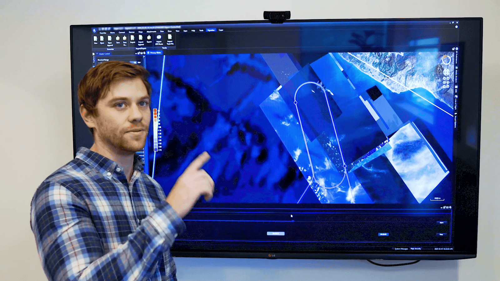 A person in a plaid shirt points at a large screen displaying satellite imagery and data visualizations.