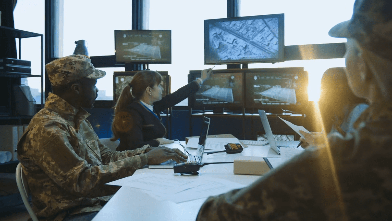 Military personnel and a woman in an office collaborate, looking at maps and screens displaying aerial images.
