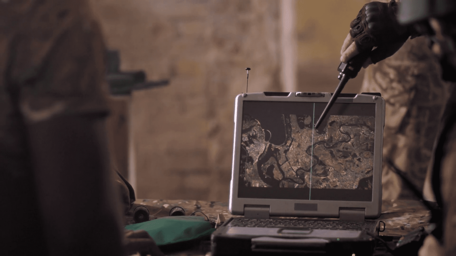 A person points with a pen at a satellite map on a laptop screen in a dimly lit room. Military equipment is visible in the background.