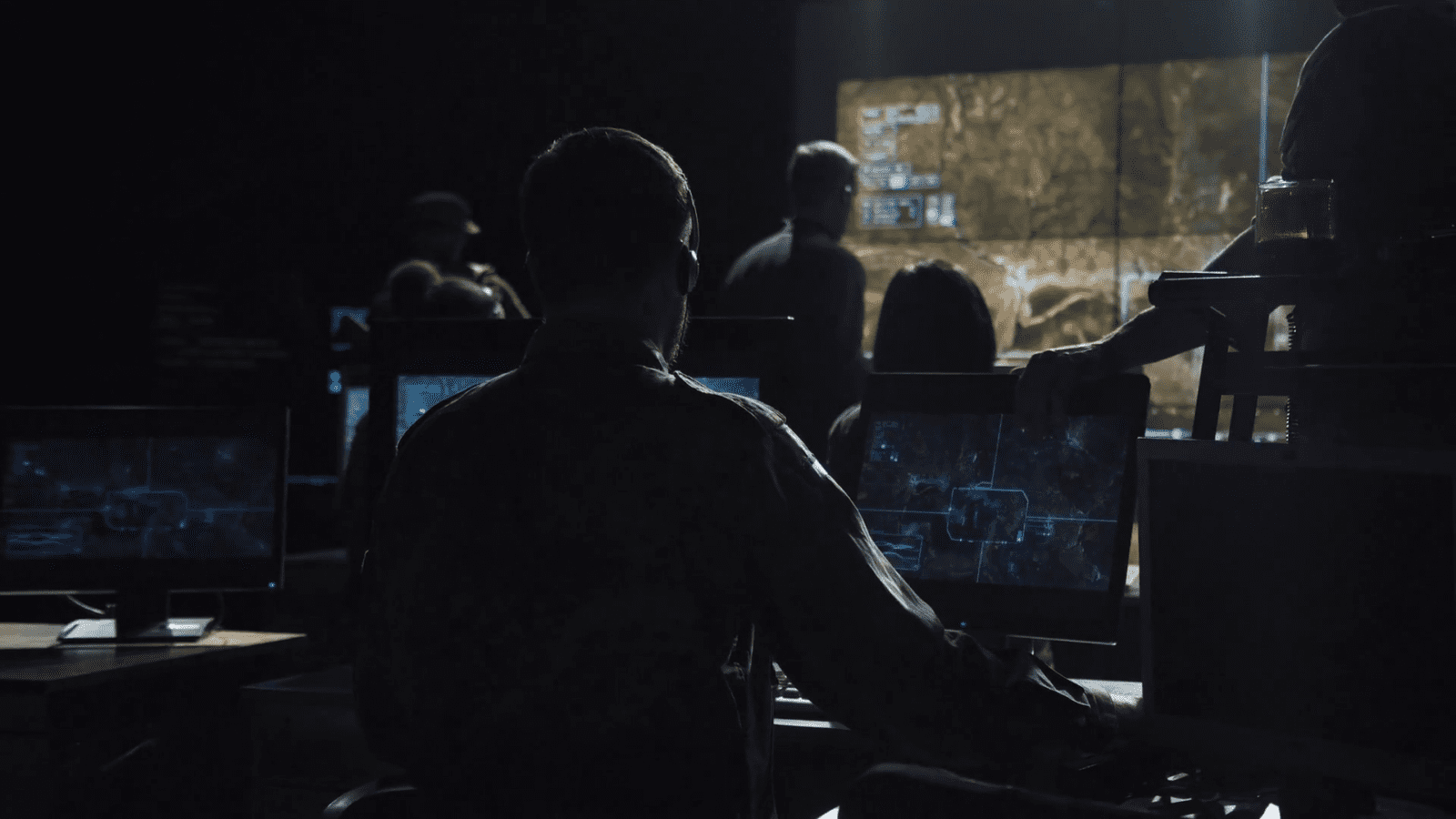 People in a dark room monitor several screens displaying maps and data, appearing focused and engaged in analysis or control operations.