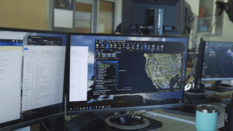 Three computer monitors showing various programs, including a map of the United States. There is a keyboard and a mug on the desk.