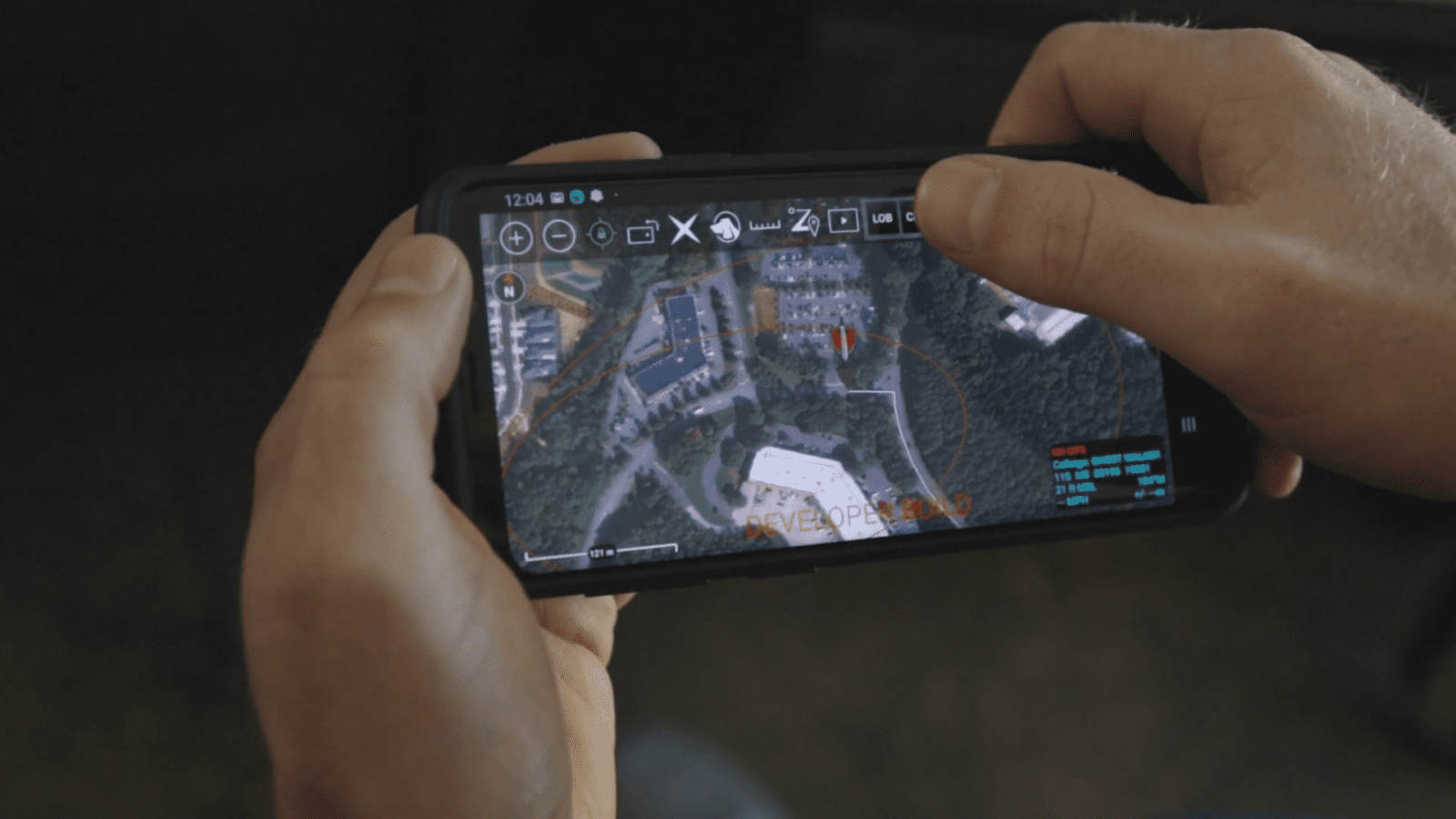 Hands holding a smartphone displaying an aerial view of a landscape with buildings, using a drone or map application interface.