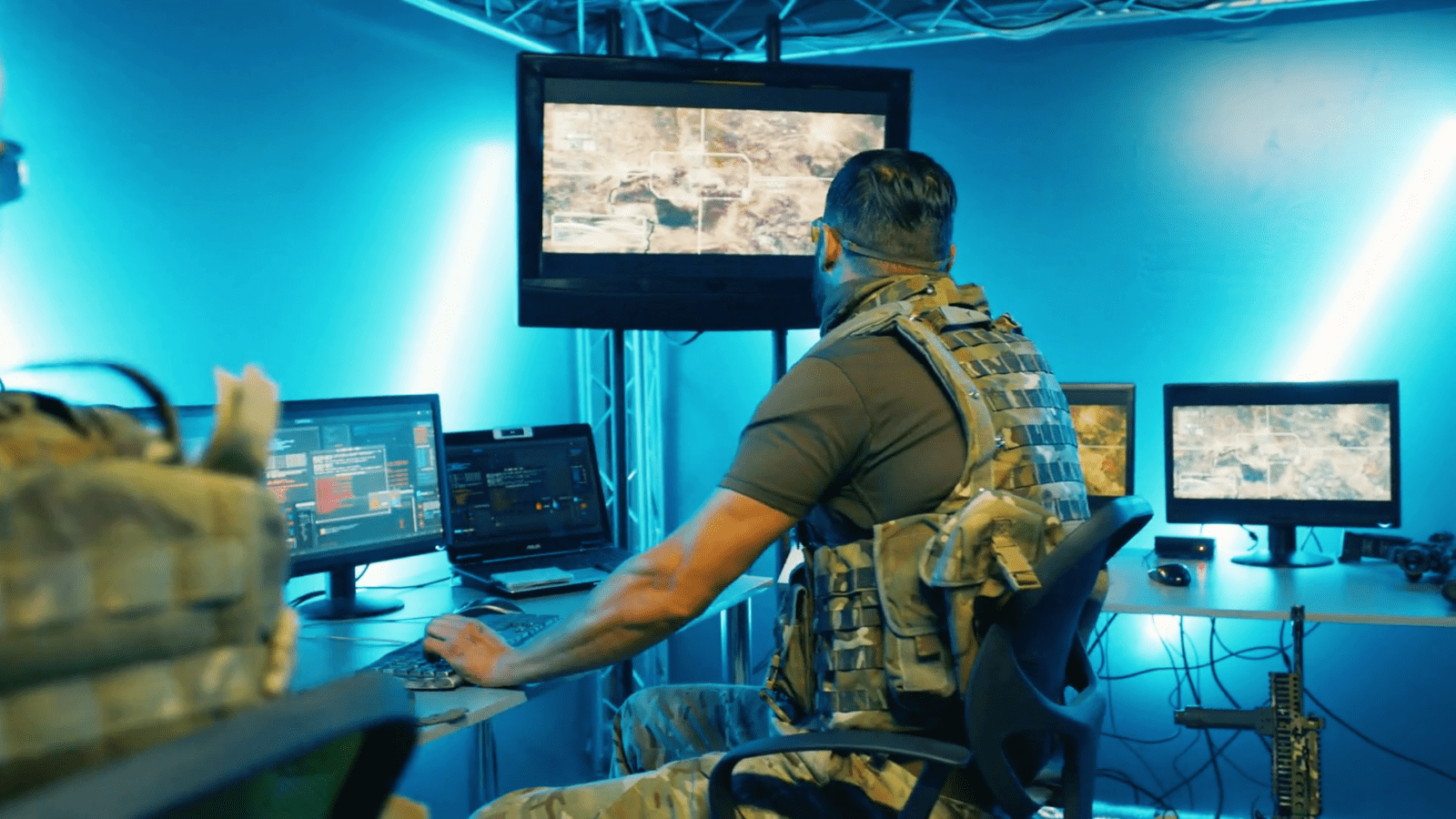 A person in military gear operates computers in a blue-lit room, focusing on screens displaying maps and data.