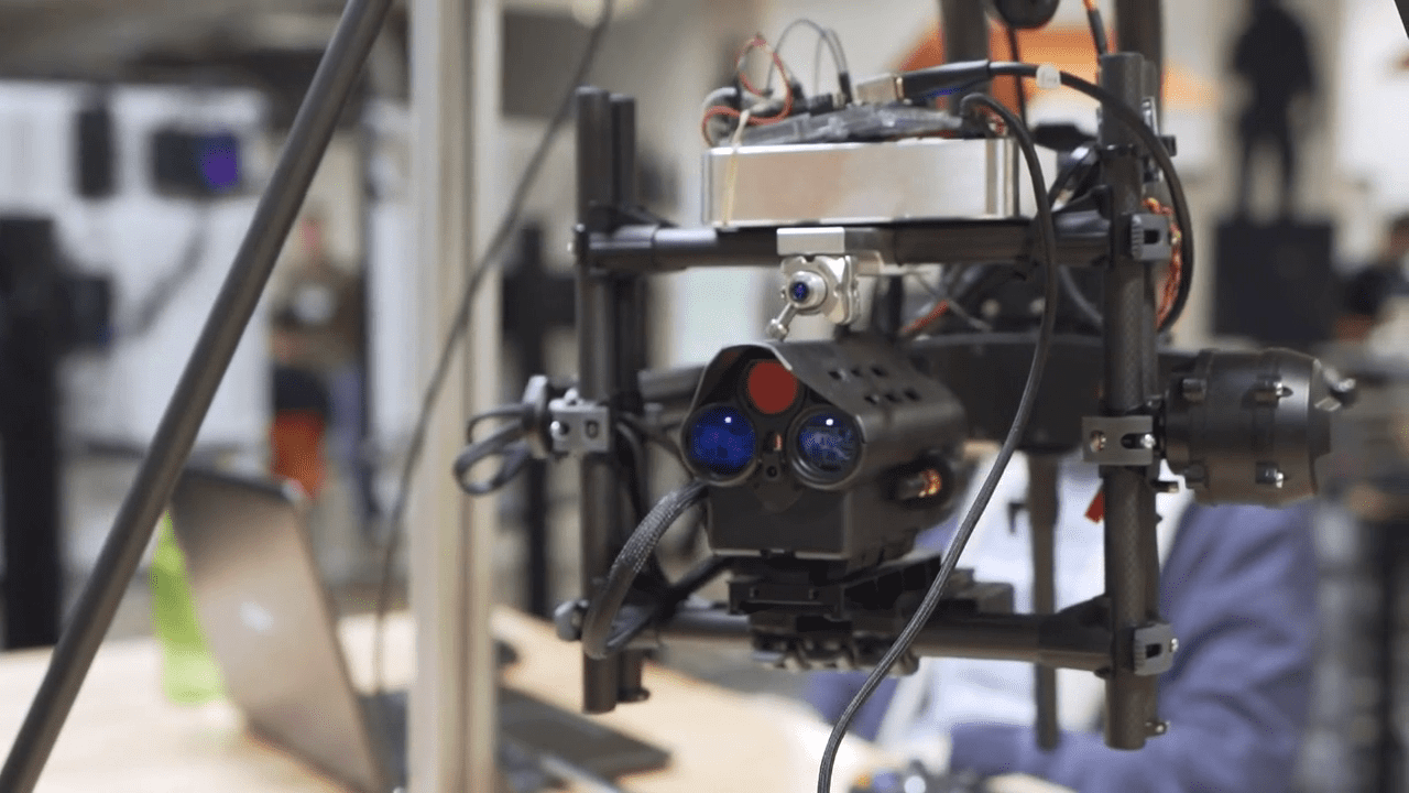 Close-up of a robotic mechanism with various wires, lenses, and sensors, in a workshop setting.
