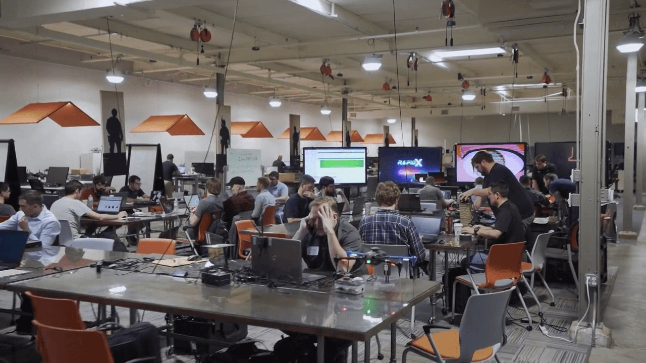 A busy office with people working at desks, multiple laptops open, large monitors displaying content, and orange-accented decor.