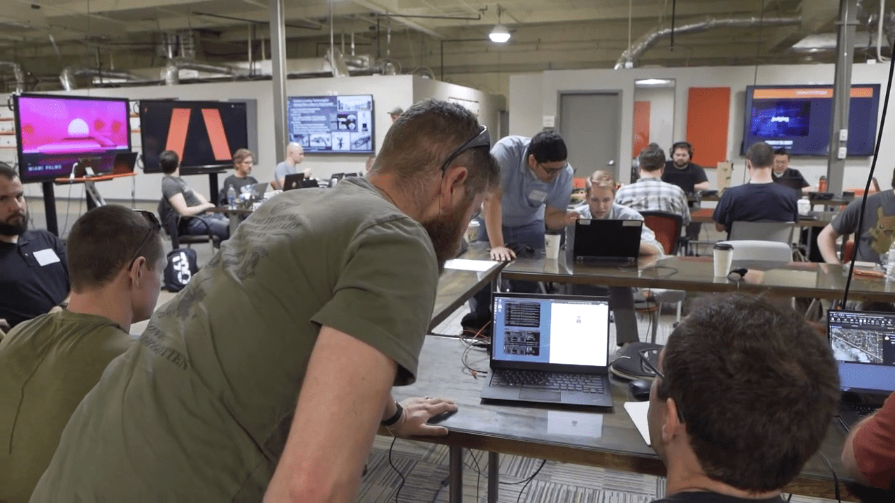 People in a classroom setting work on laptops and collaborate. A presentation is displayed on screens at the front.