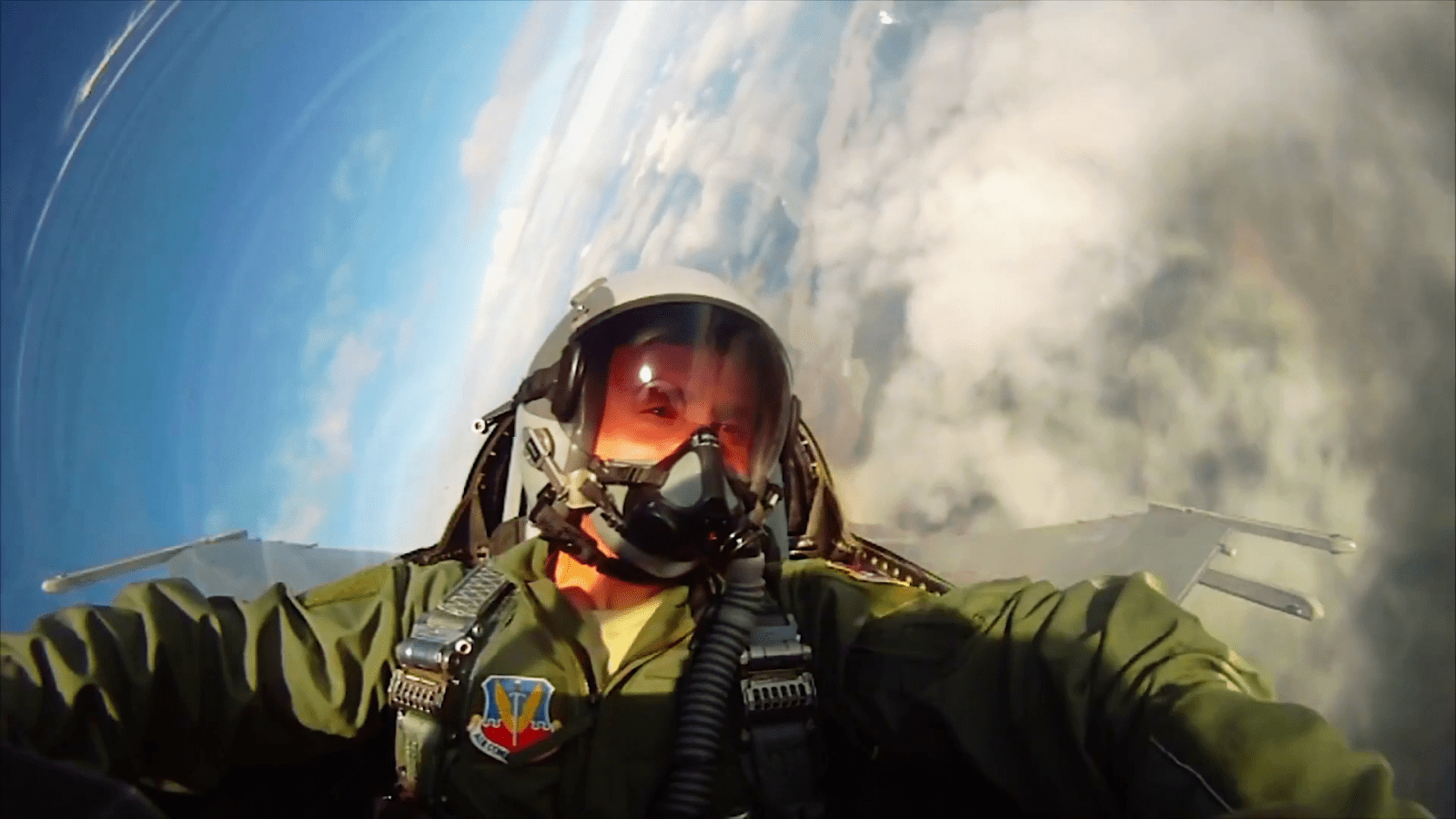 A pilot in a flight suit and helmet flies a jet, with clouds visible in the background.