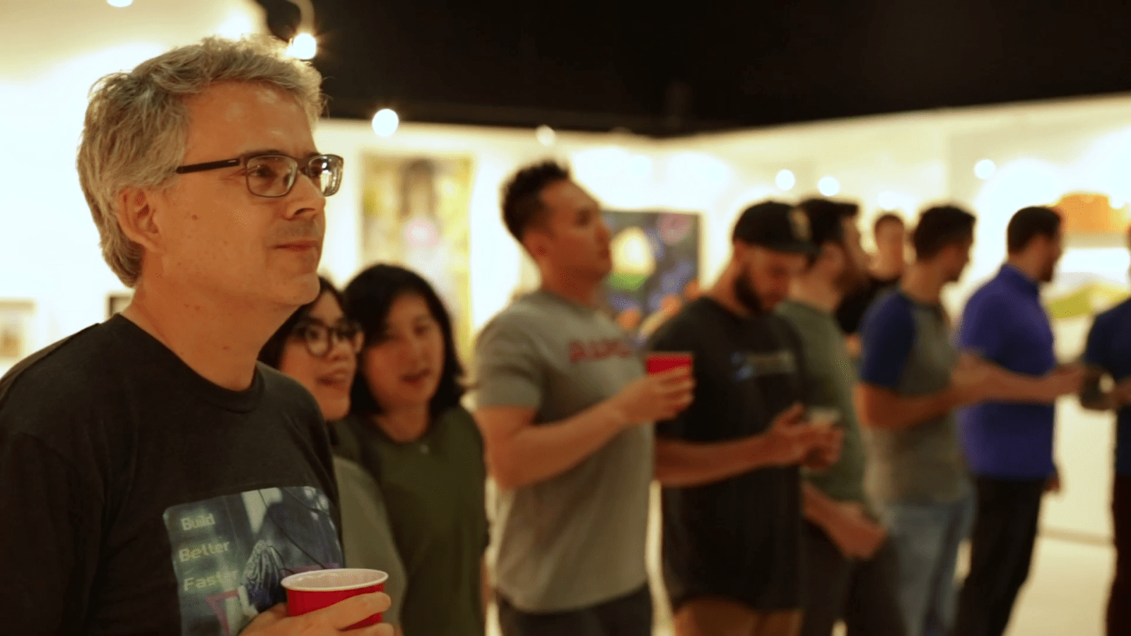 People standing together at a social event, holding red cups and engaging in conversation. Artworks are visible in the background.