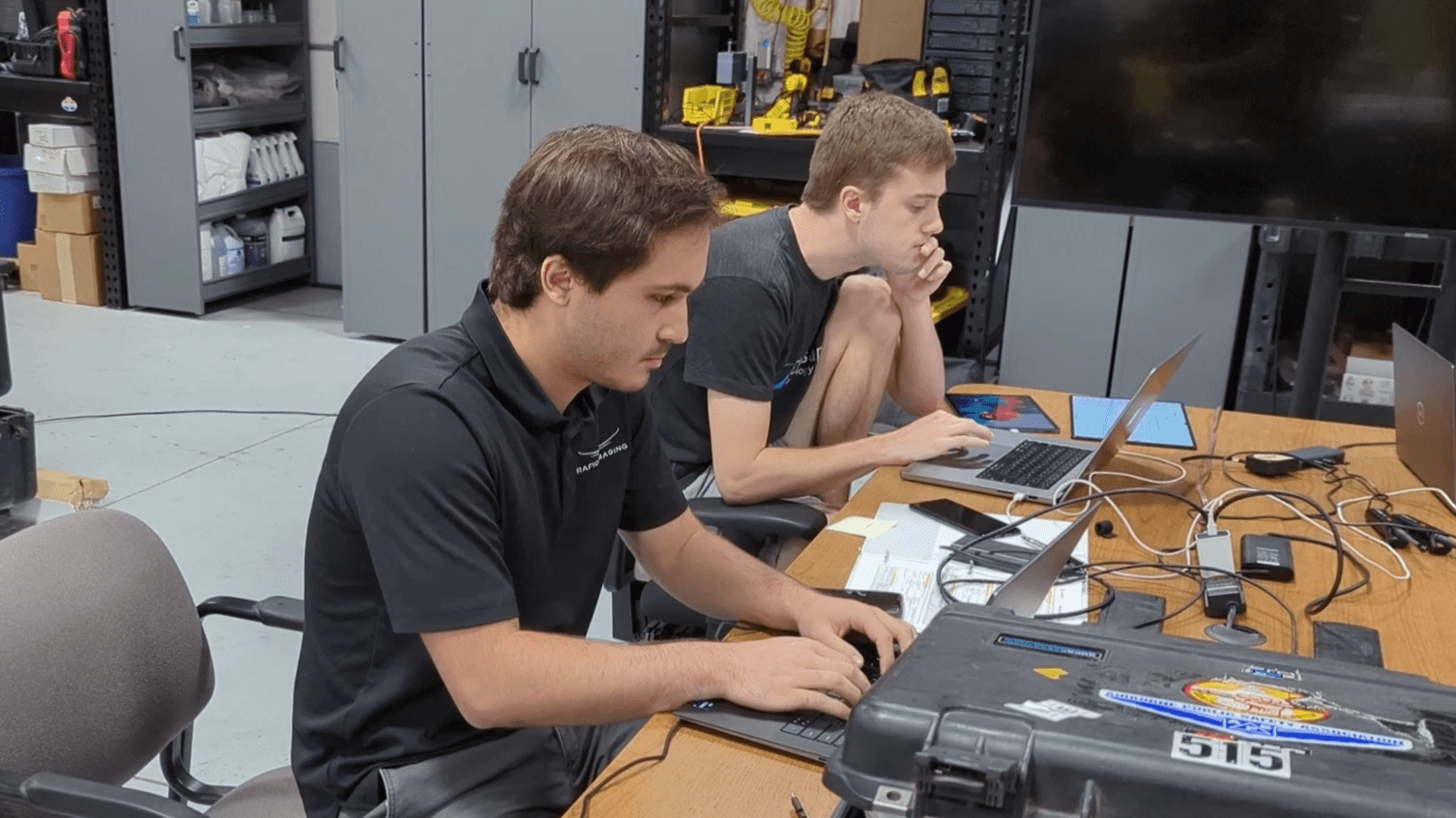 Two people sit at a table in a workshop, using laptops. The table is cluttered with equipment and a black case. Shelves with tools and a large screen are in the background.