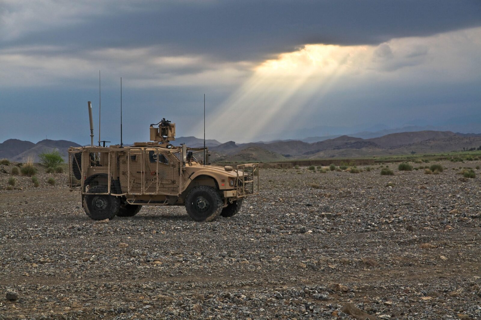 A military vehicle is parked on a rocky terrain under a cloudy sky with sun rays breaking through.