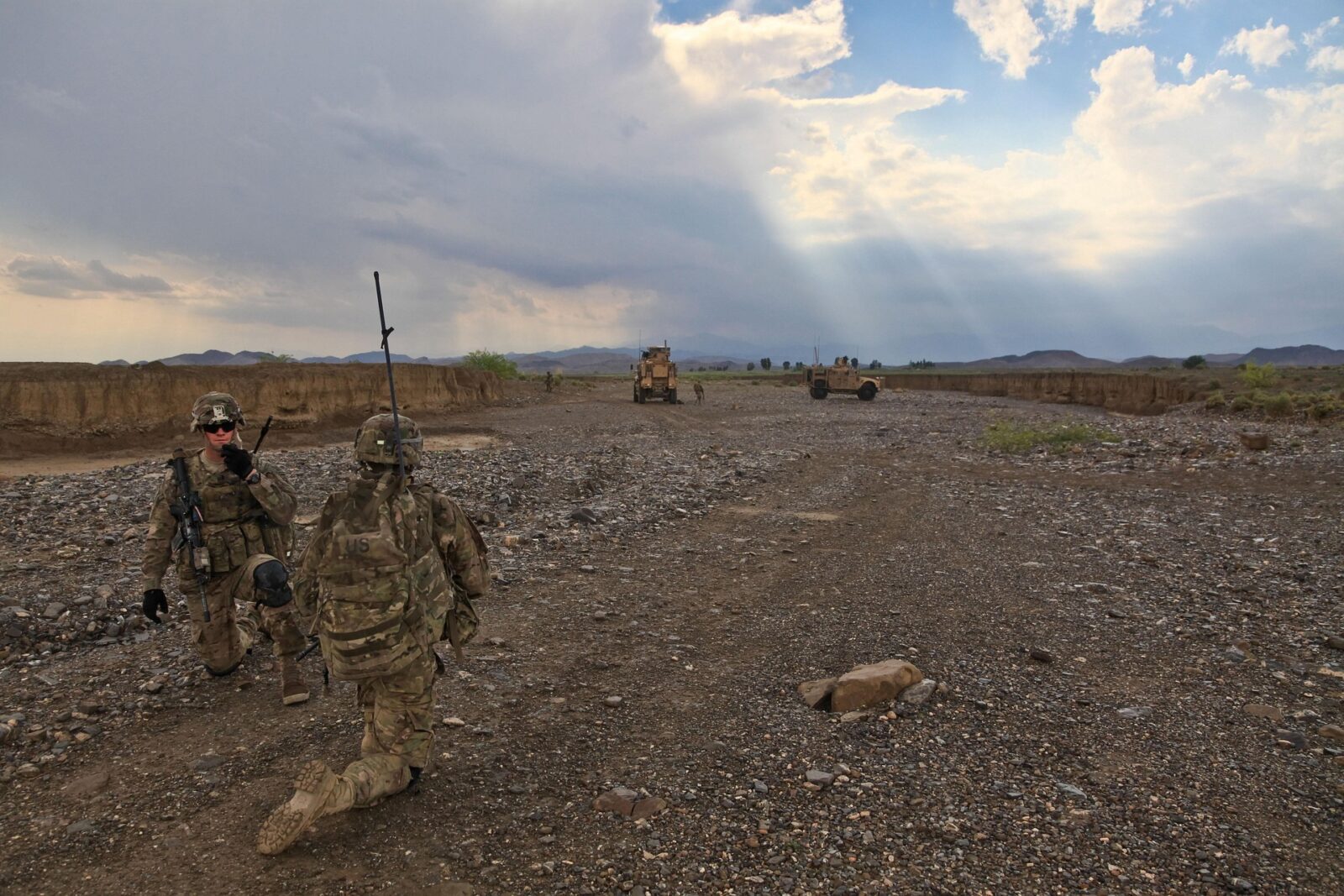Two soldiers in combat gear communicate using a radio in a rocky landscape, with military vehicles and a cloudy sky in the background.