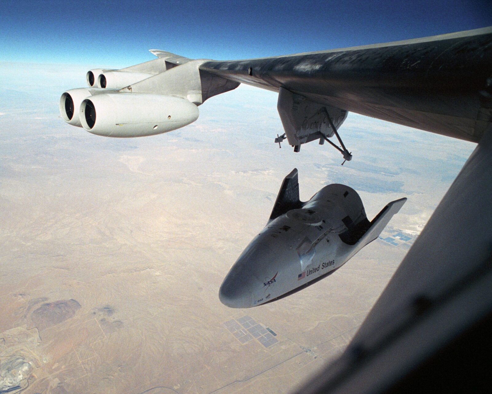 Spacecraft attached under the wing of a large plane in flight over a desert landscape.