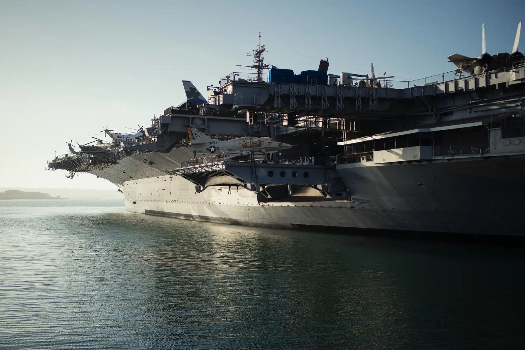 A large aircraft carrier is docked at a harbor, with various military aircraft visible on its deck under a clear sky.