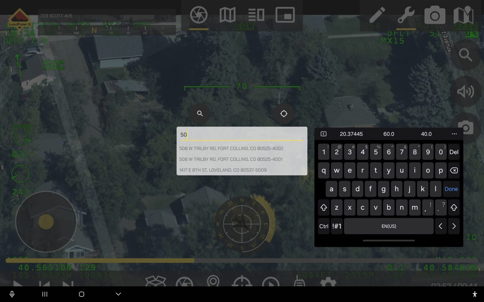 Aerial view on a screen showing a satellite image of houses with overlaid coordinates and numbers. A keyboard and a search box with addresses are visible on the screen.