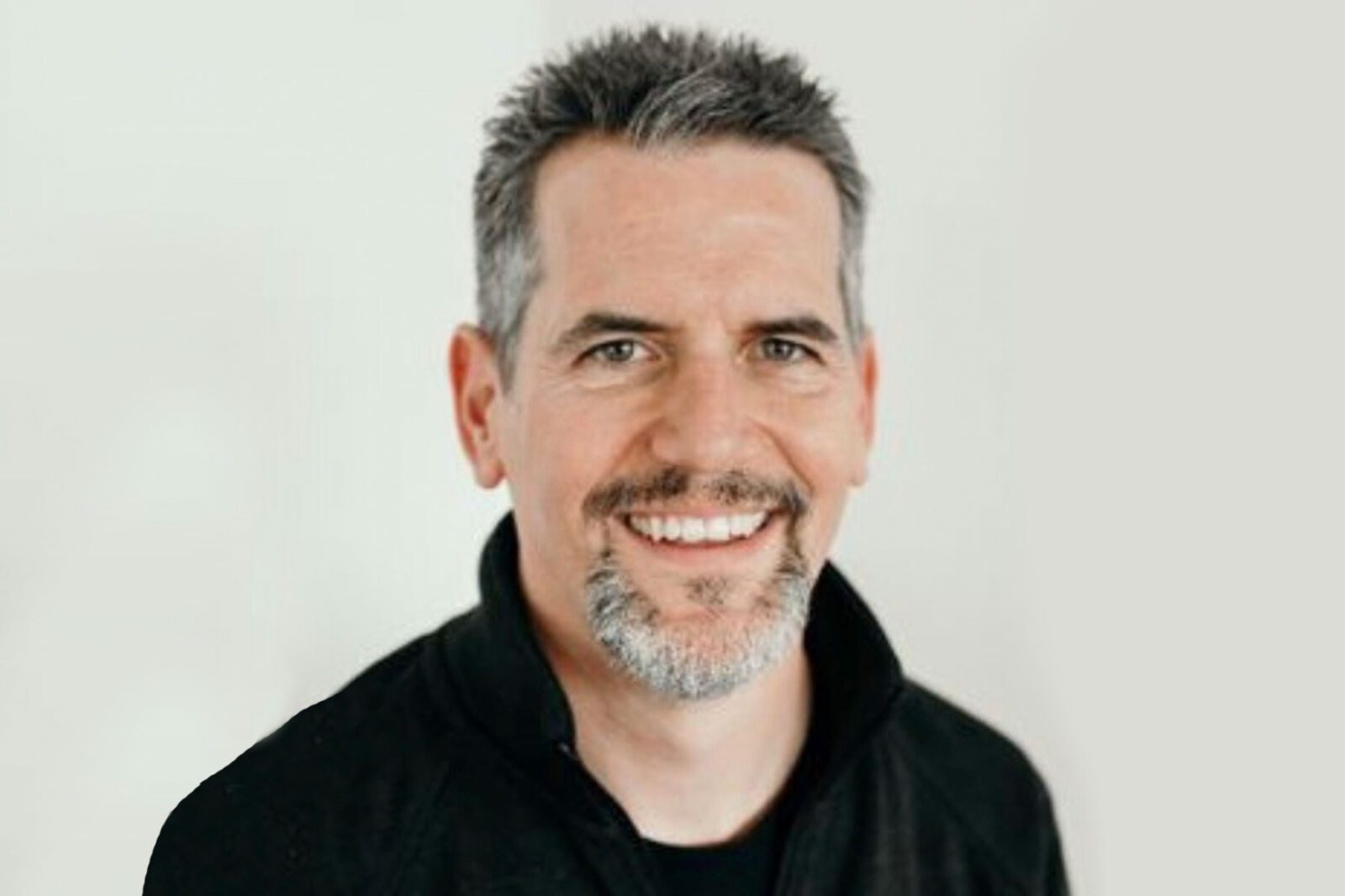 Smiling man with short gray hair and a goatee, wearing a dark jacket, against a plain background.