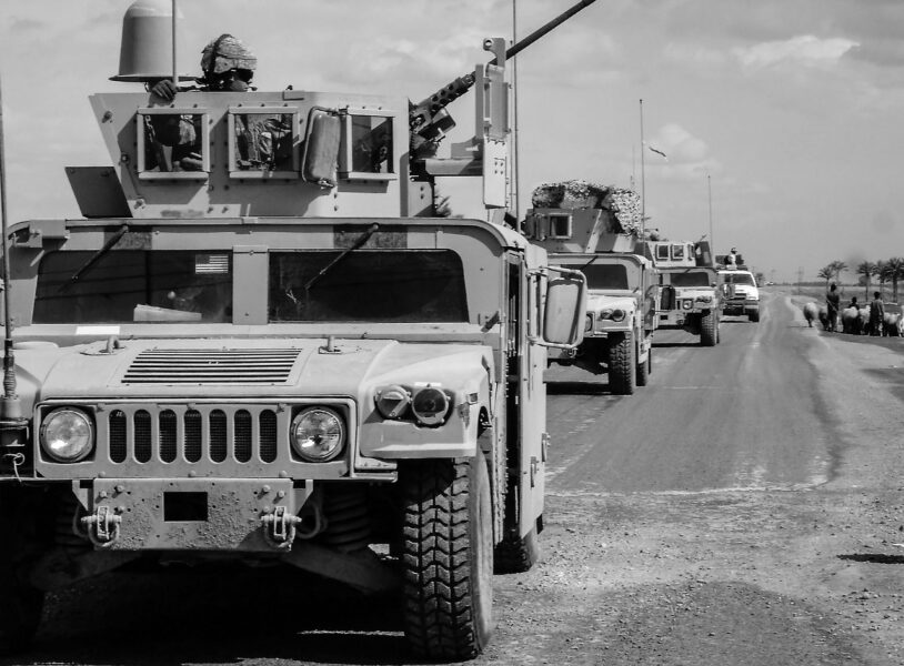 A convoy of military vehicles travels on a paved road in a rural area under a partly cloudy sky.