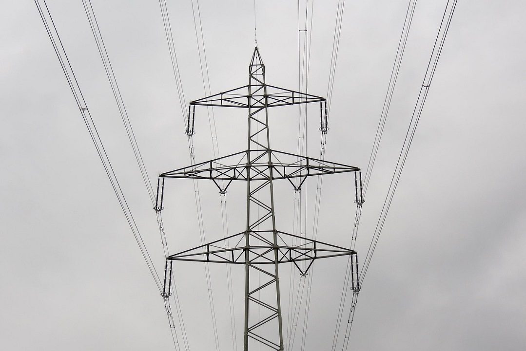 A metal electricity pylon with multiple power lines extends upwards against an overcast sky.