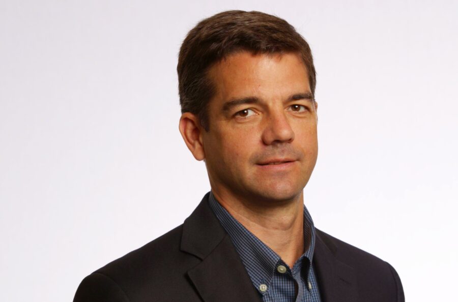 A man with short brown hair wearing a dark suit jacket and blue shirt against a plain white background.