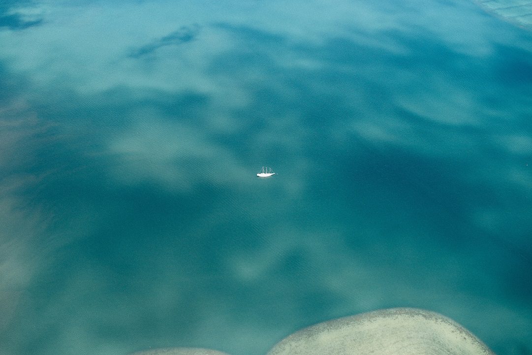 Aerial view of a small white sailboat on calm, turquoise water, surrounded by subtle cloud reflections.