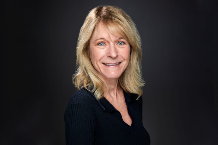 A woman with shoulder-length blonde hair and a dark top smiles at the camera against a dark background.