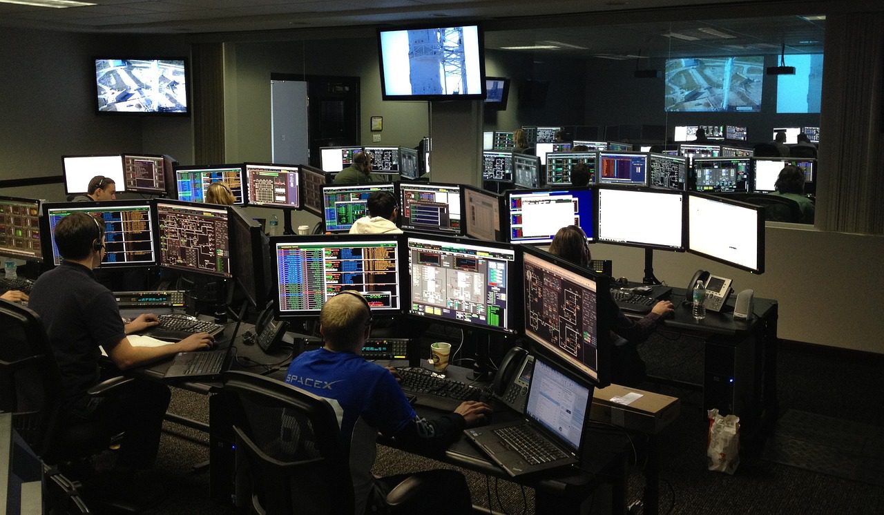 A group of people working at computer stations with multiple monitors in a dimly lit control room.
