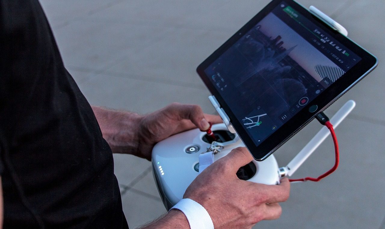 Person operating a drone controller, with a tablet displaying aerial footage and navigation controls.