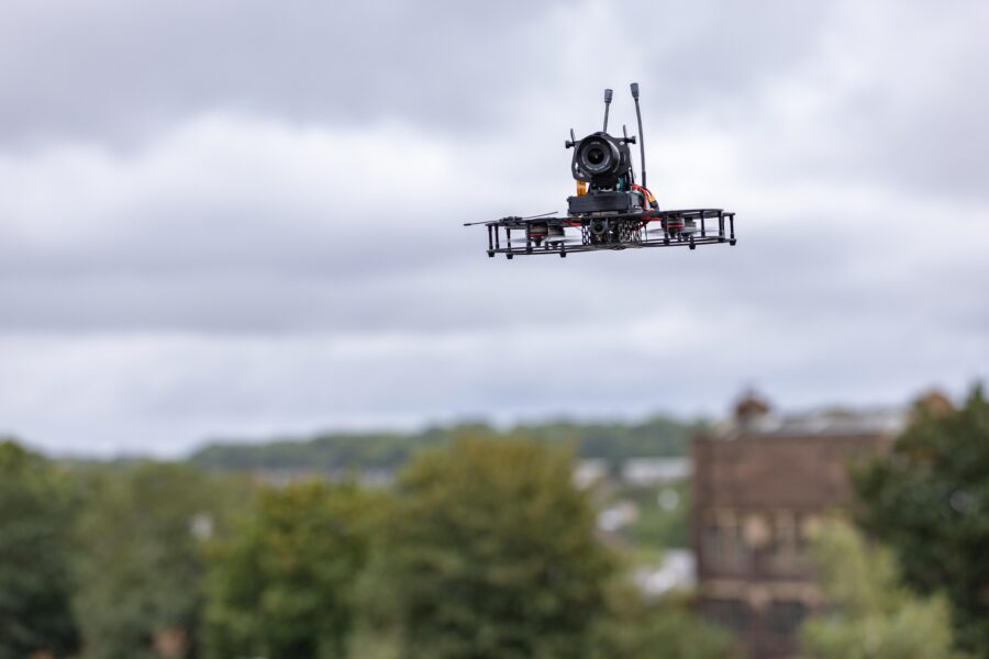 A small drone with a camera flies outdoors against a cloudy sky, with blurred trees and buildings in the background.