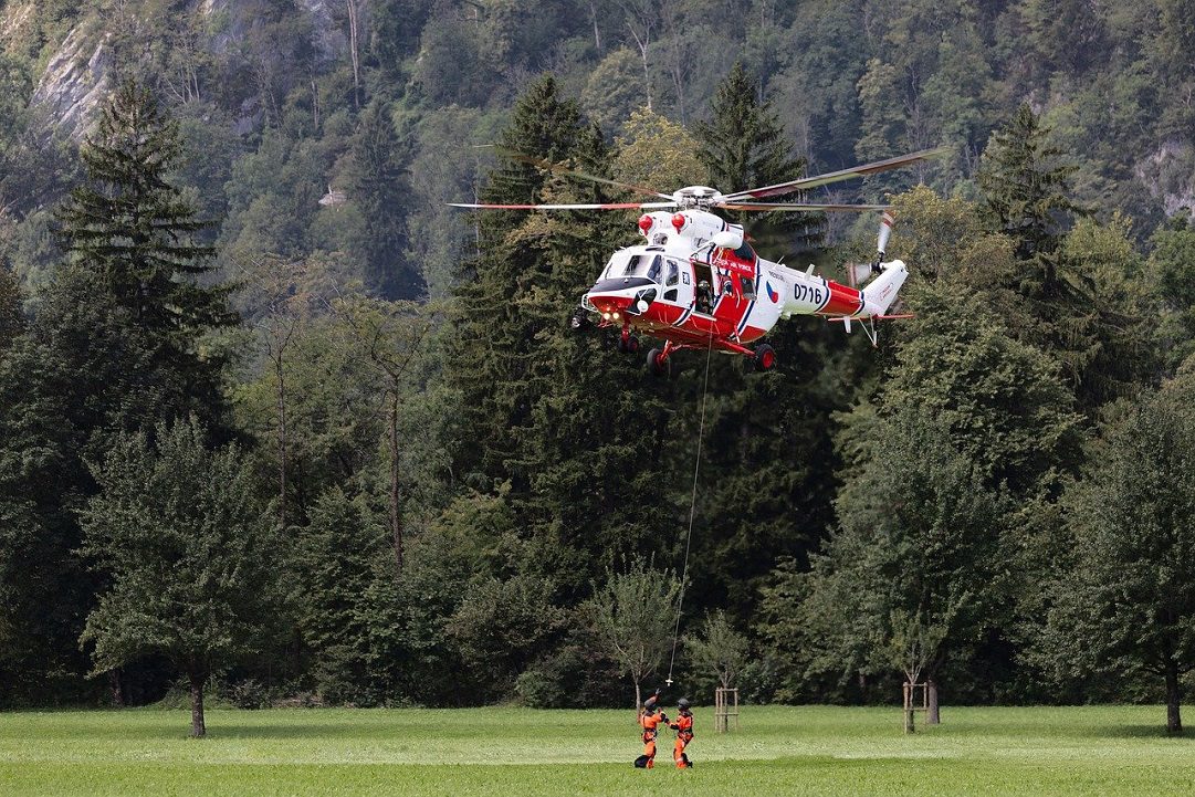 A red and white helicopter hovers over a grassy field, lowering two rescue workers using a cable. Forested hills are in the background.
