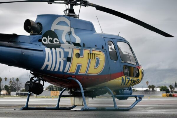 Blue and yellow news helicopter on the ground at an airfield with cloudy skies and distant mountains in the background.