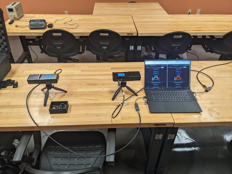 A wooden table with electronic devices, two webcams, and a laptop displaying graphs. Black chairs are arranged around the table in a room setting.