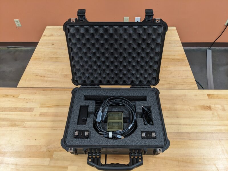 Open black hard case with foam padding containing electronic equipment, cables, and small accessories, placed on a wooden table.