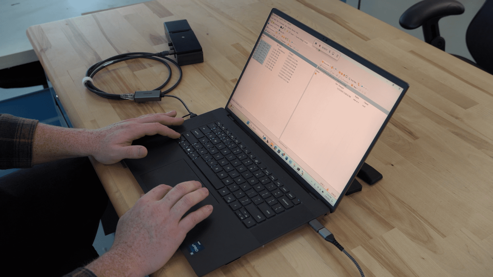 Person typing on a laptop with a split screen displaying text files. A USB cable and black adapter rest on the wooden table nearby.