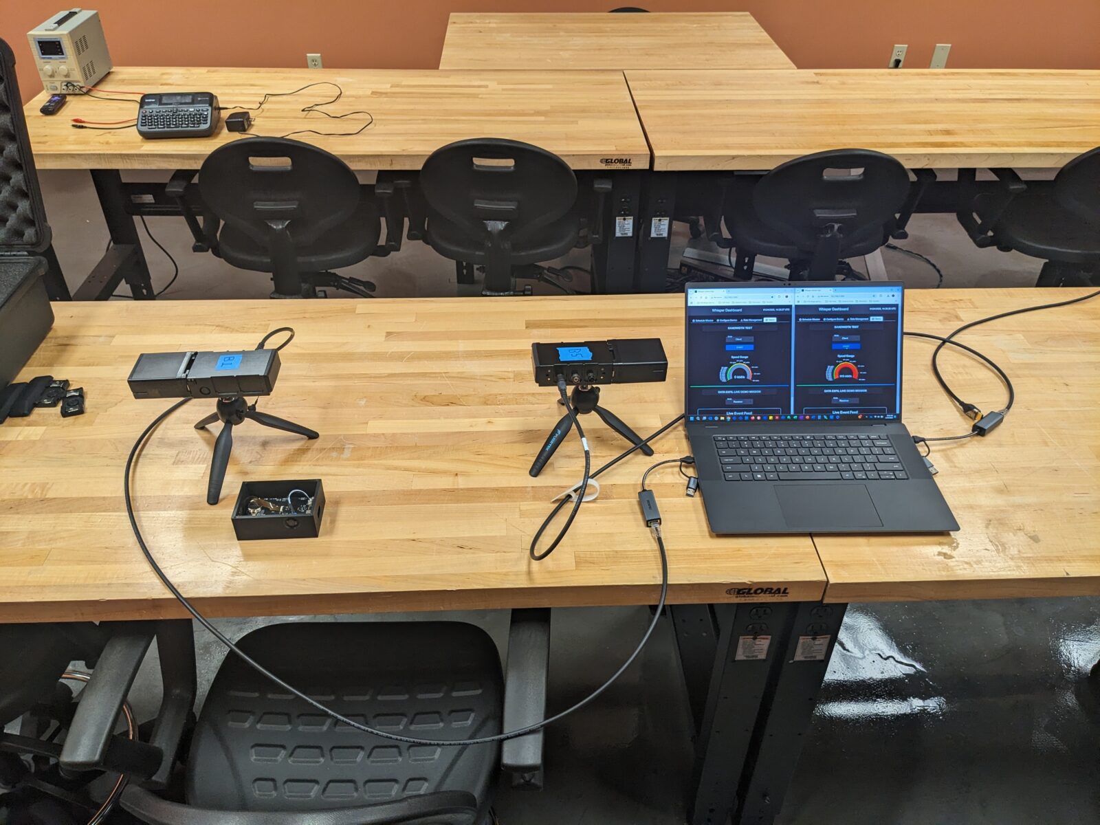 A wooden table with electronic devices, two webcams, and a laptop displaying graphs. Black chairs are arranged around the table in a room setting.