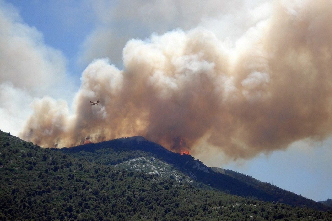 Aerial view of a forest fire on a mountain, with thick smoke rising. An airplane flies nearby against a blue sky.
