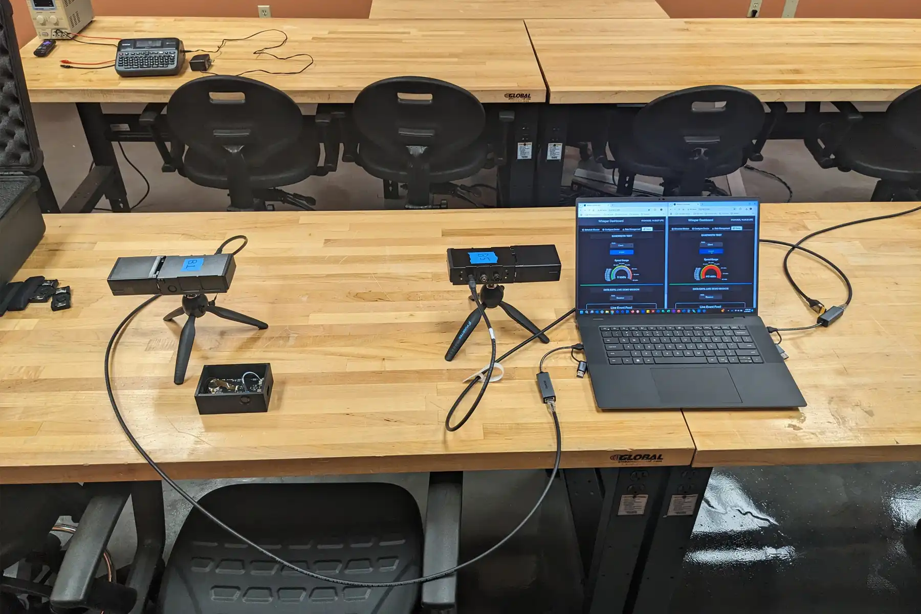 Workspace with wooden tables, two cameras on tripods, a laptop showing data, and several chairs.