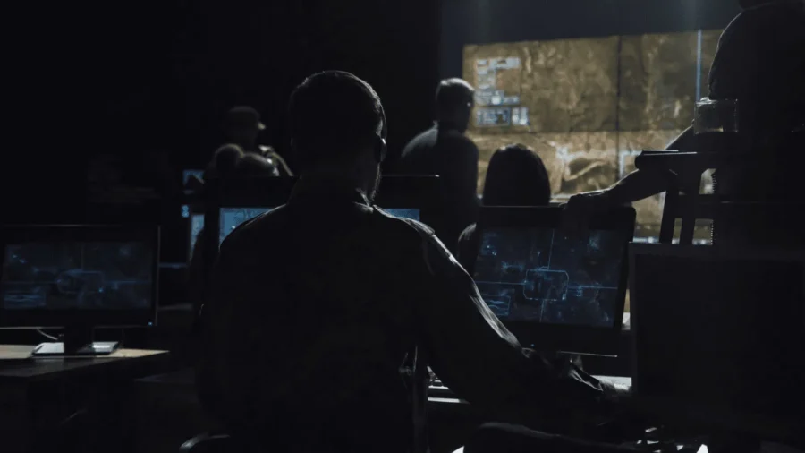 A dimly lit control room with people working on multiple computer screens, analyzing data from large digital displays on the wall.